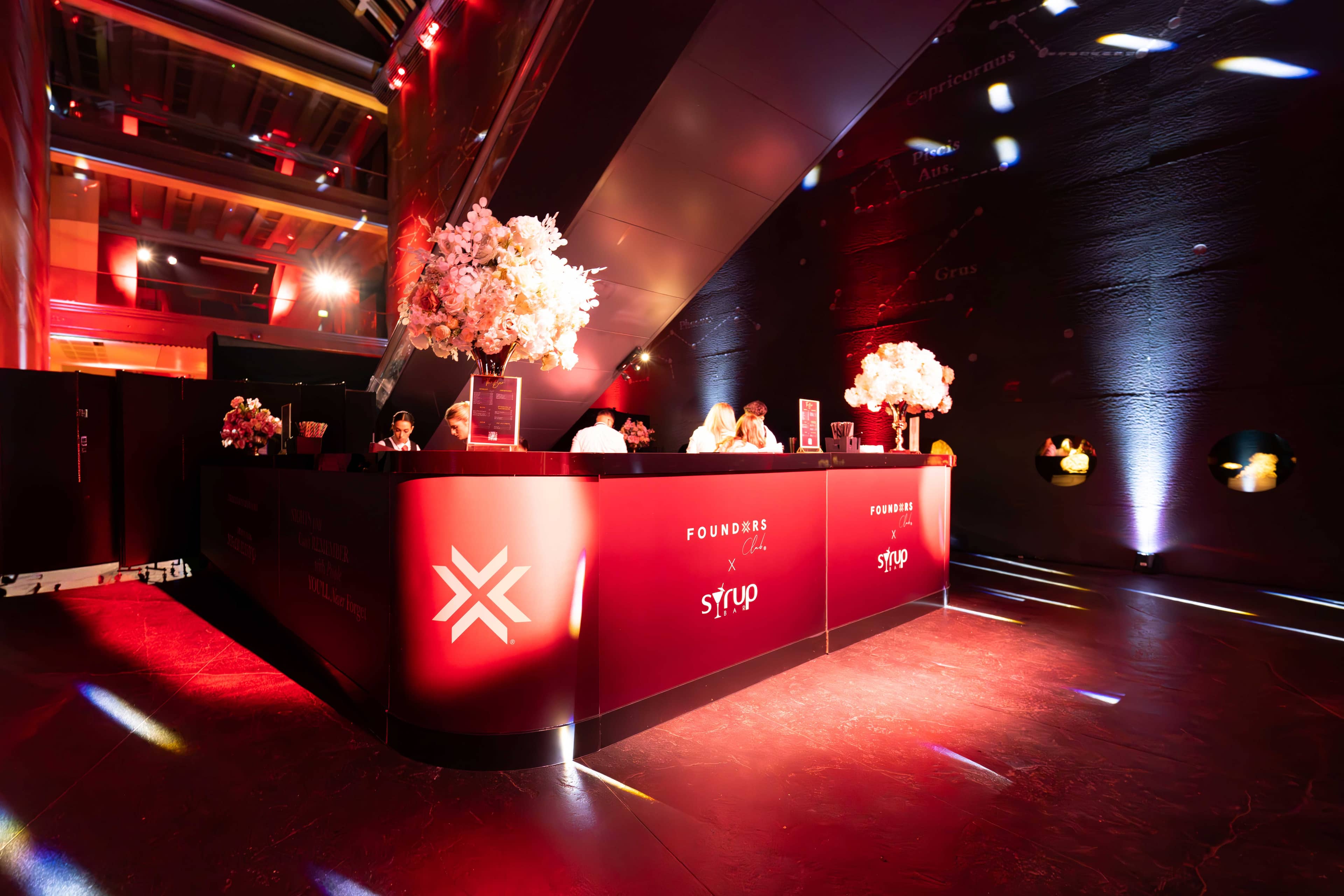 The red reception desk with white florals under red and blue uplighting at the FOUNDXRS Club black tie party in Earth Hall, by Kudos AV.