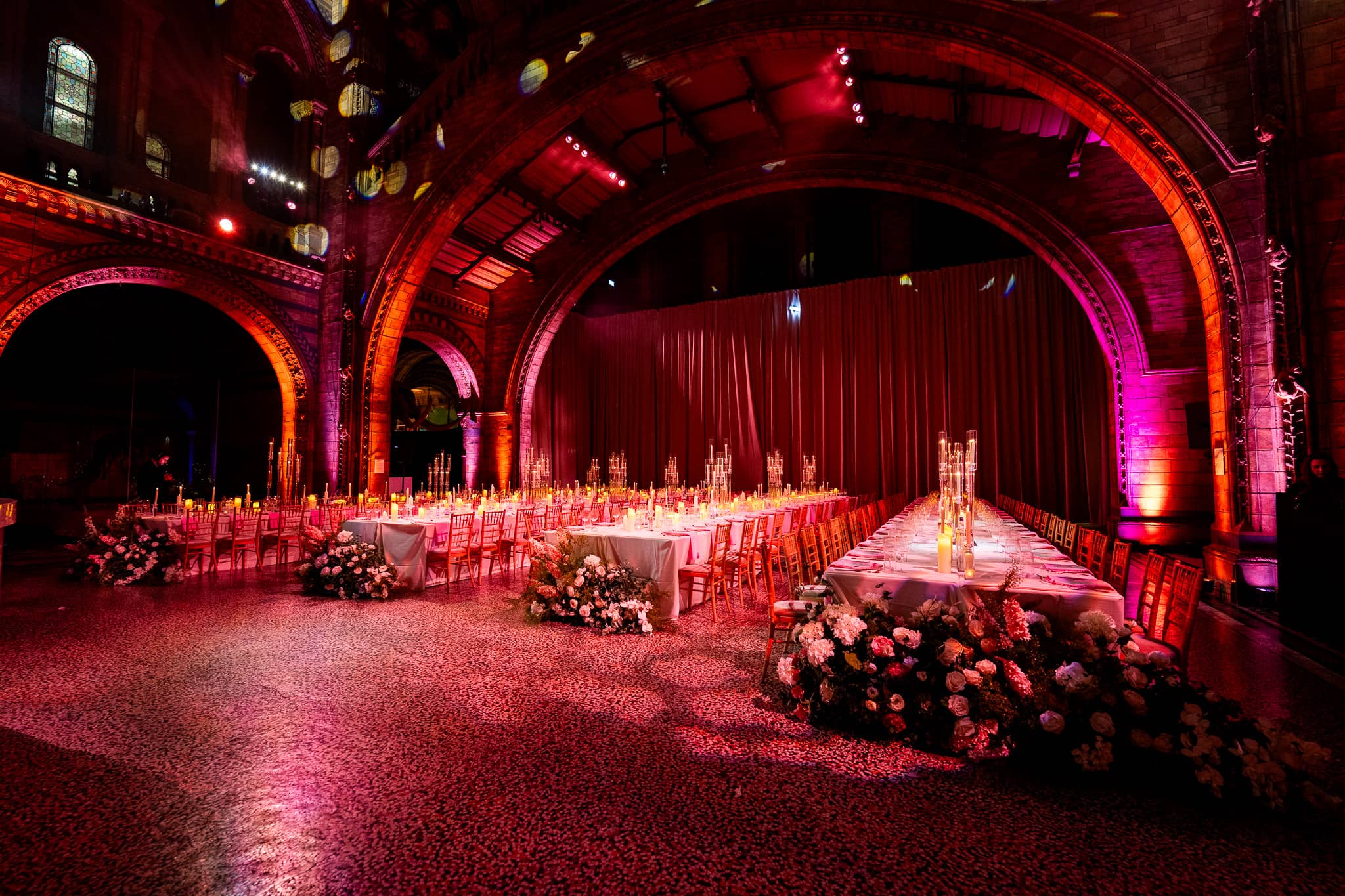 White floral centrepieces on long dinner tables in Hintze Hall under red uplighting, a Natural History Museum wedding lit by Kudos AV.