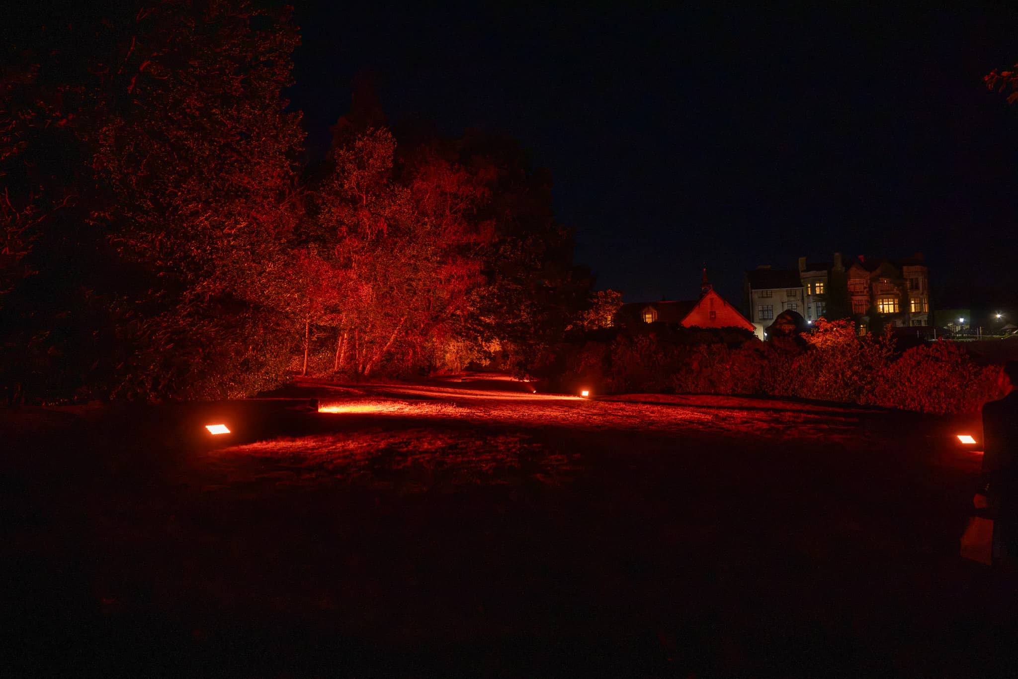 The house at Pennyhill Park in the background with the trees and lawns in front of it uplit red for a marquee event, Surrey, by Kudos AV