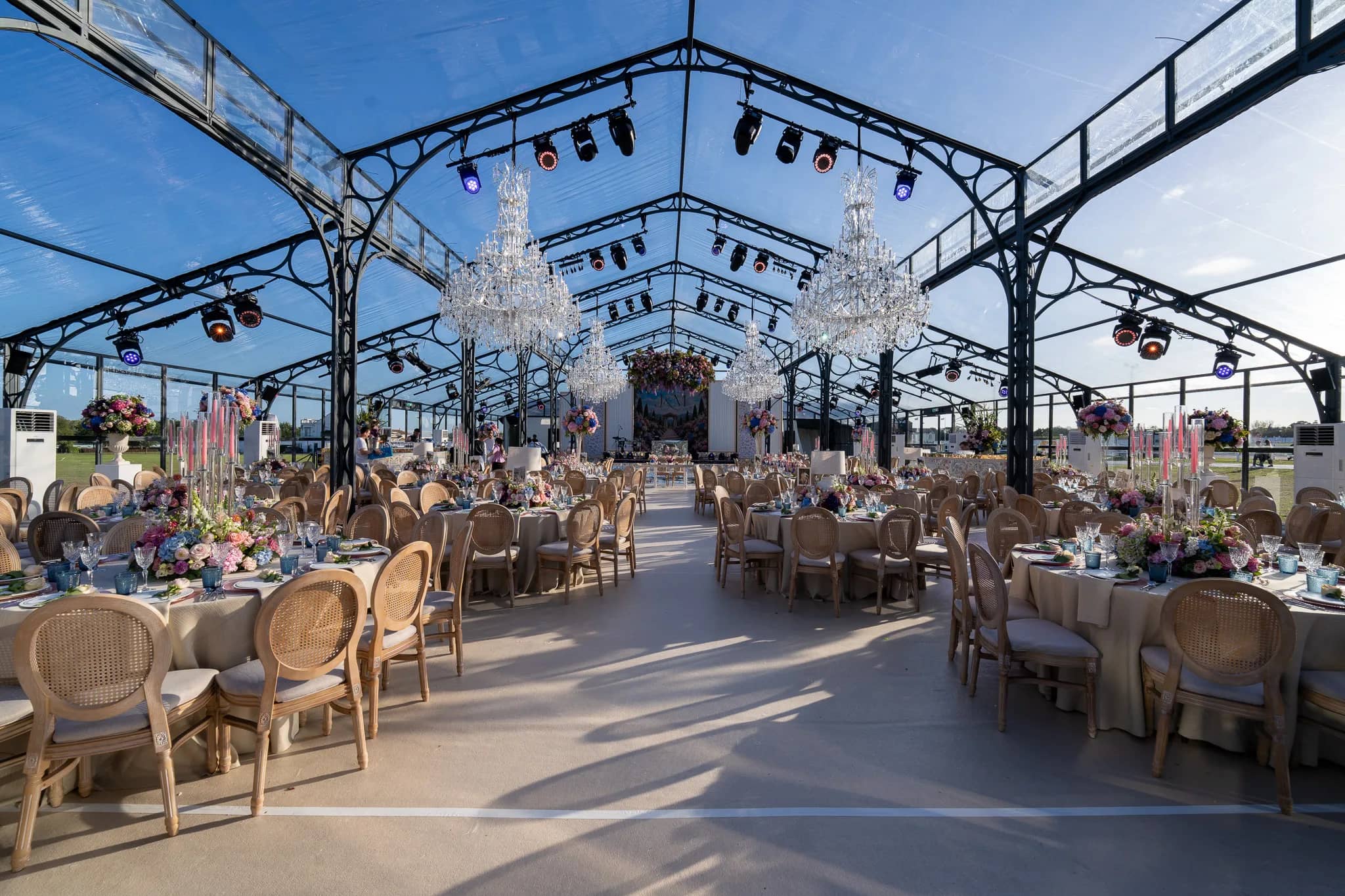 Round tables with wildflower centrepieces and taper candles down the length of a glass marquee, two crystal chandeliers and moving lights on the frame above, Warwickshire, by Kudos AV