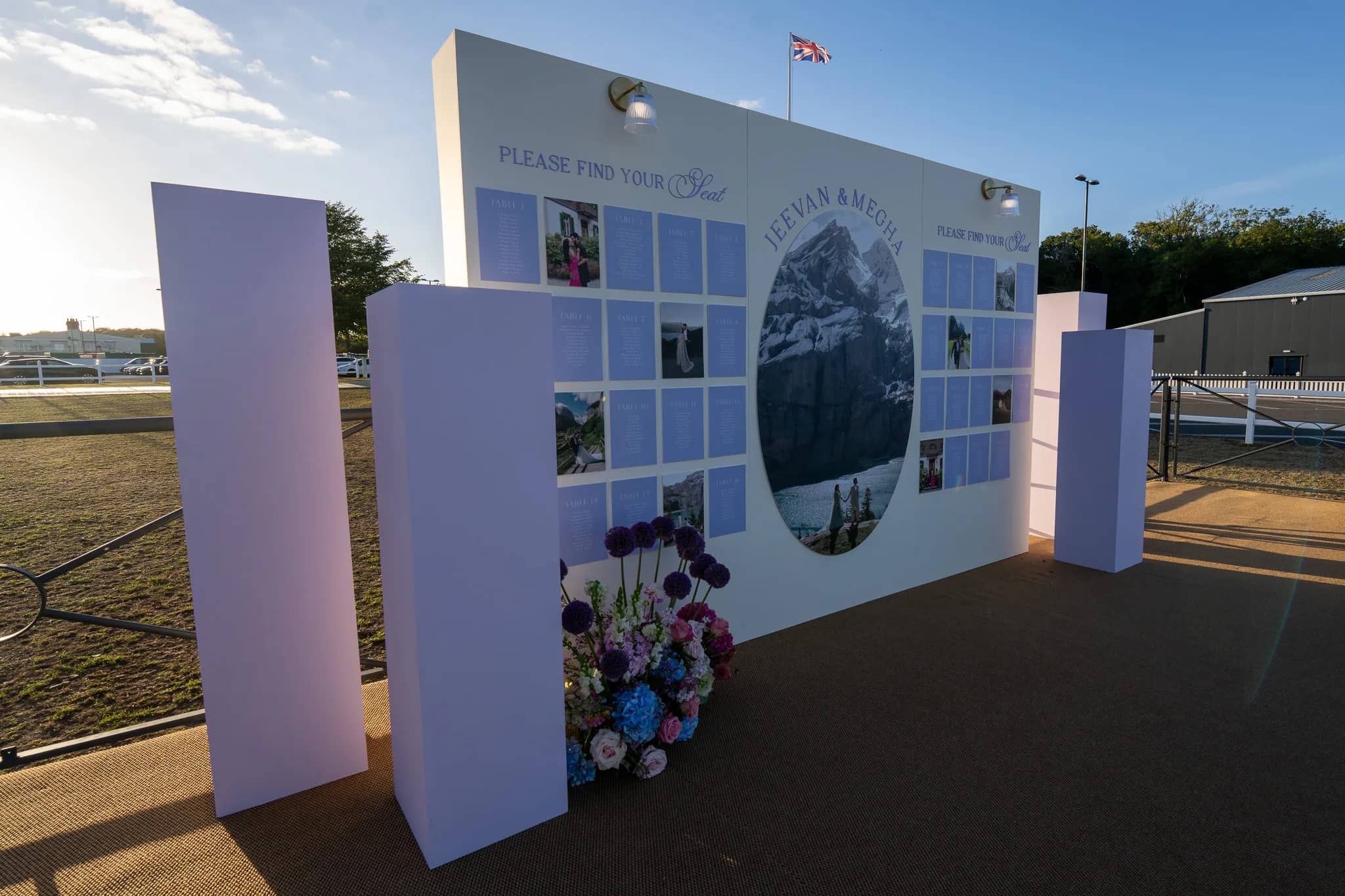 A seating plan wall with a photograph of the couple in the Alps, lilac plinths and a wildflower arrangement at its foot, on parkland in Warwickshire, by Kudos AV