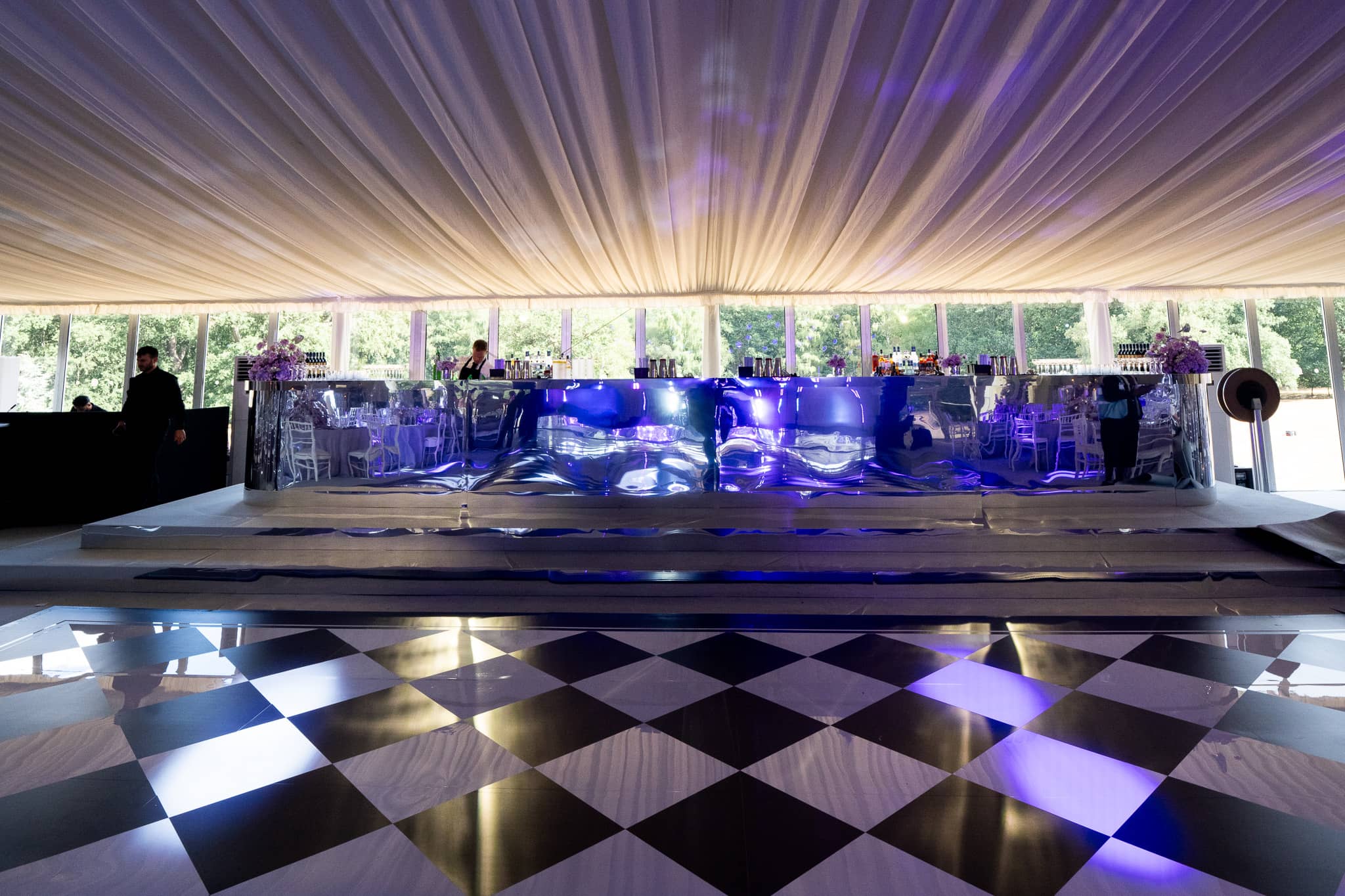 A raised bar printed in a mirror finish and lit blue, with a chequerboard mirror dancefloor in front of it and purple uplighting along the marquee walls, Pennyhill Park, Surrey, by Kudos AV