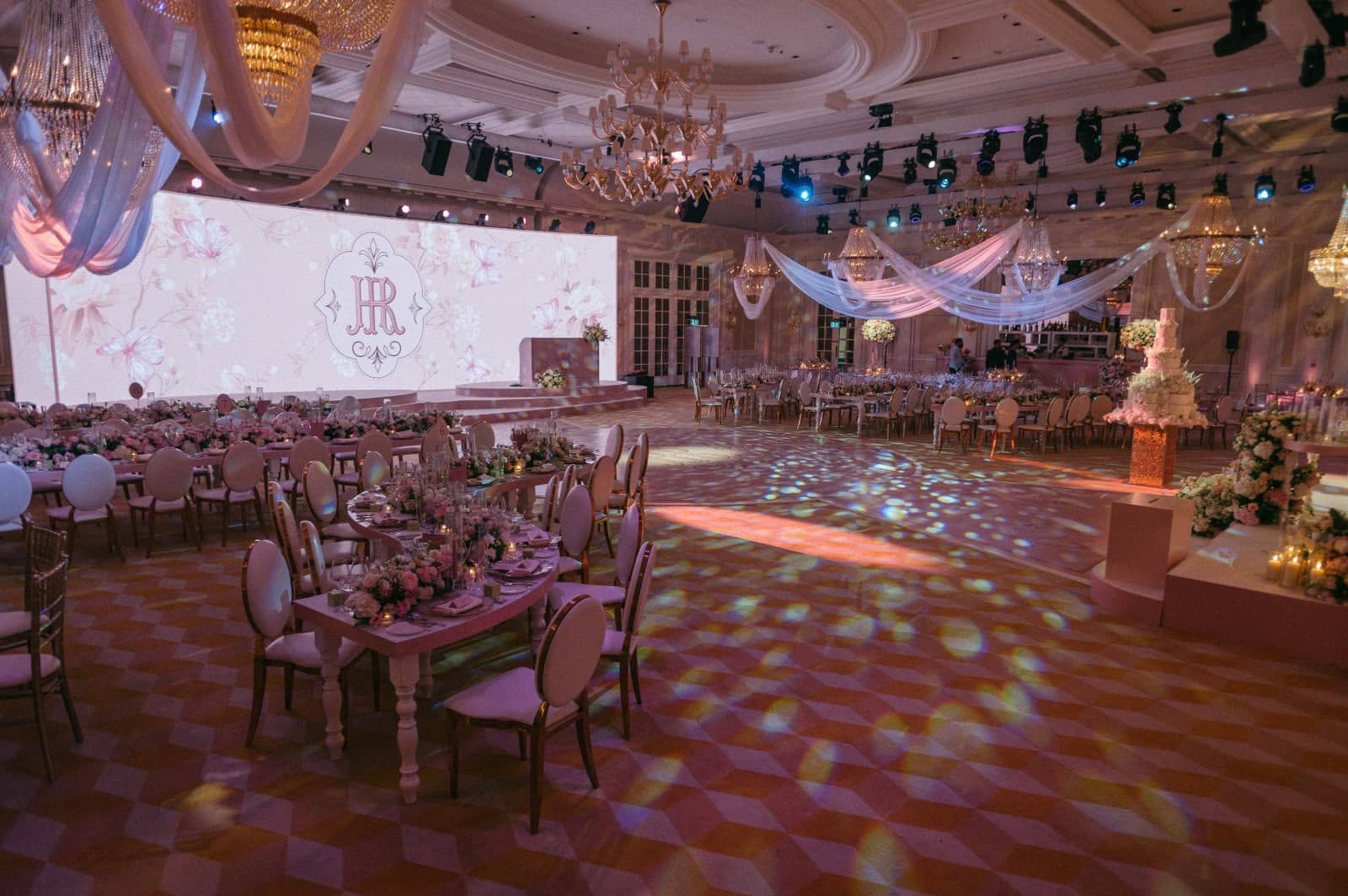 Patterned light falls across the ballroom dance floor beside guest tables, facing a pink floral LED screen and cake display at Raffles London.