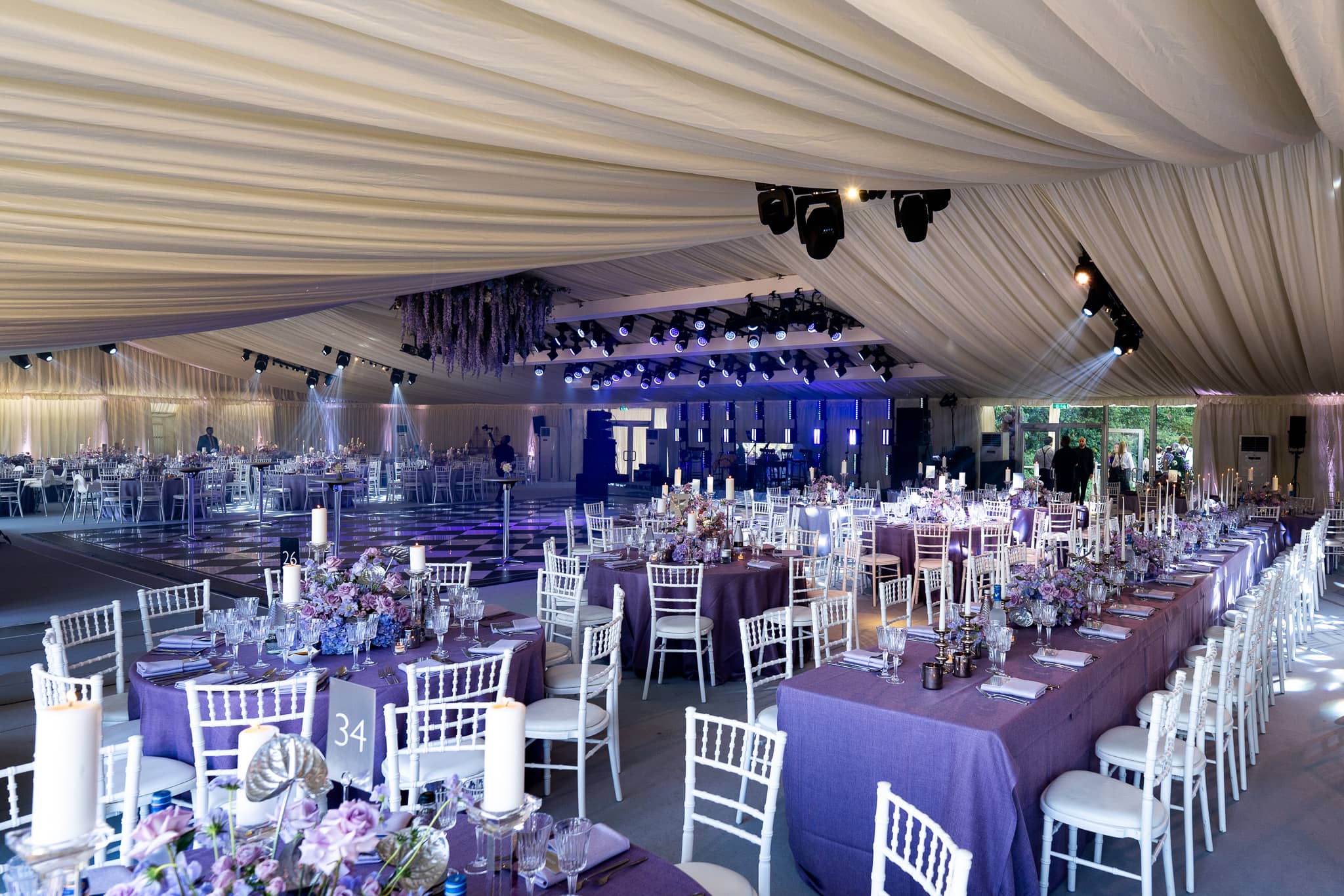The white draped interior of a luxury marquee in purple uplighting, round tables dressed with florals for 500 guests, Pennyhill Park, Surrey, by Kudos AV