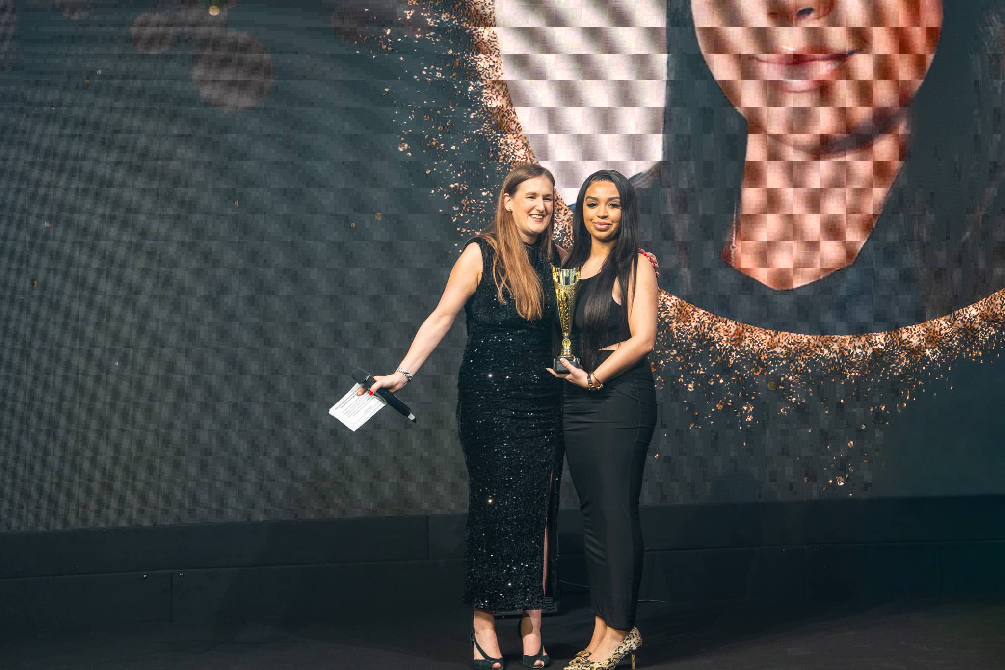 Two people pose with an award in front of a winner portrait on the MFS LED screen