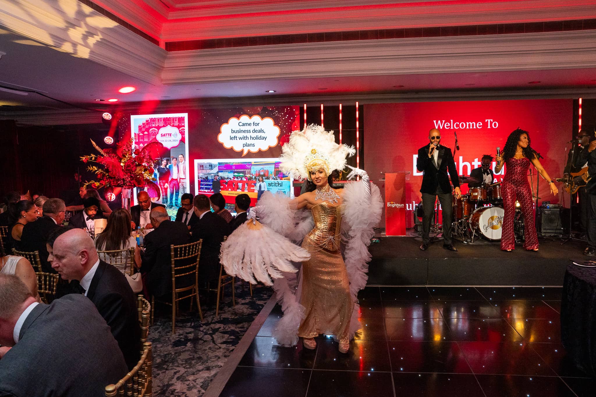 Showgirl performing beside dinner guests, with Goldn Global on stage behind, at Brightsun Travel’s 40th anniversary at InterContinental London Park Lane.