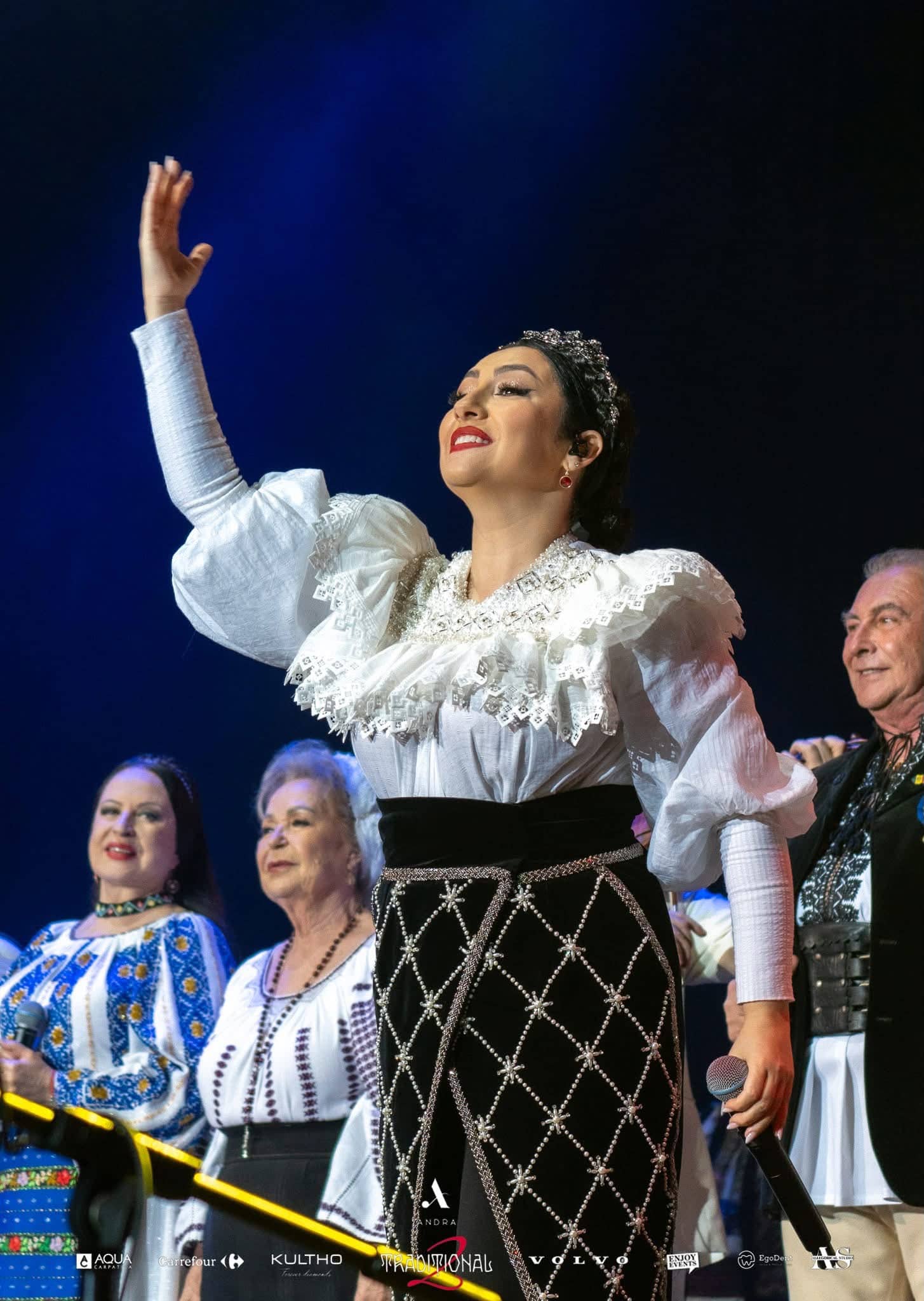 Andra in a white ruffled blouse raising her hand to the crowd, guest singers in traditional dress beside her, Andra Traditional 2 at OVO Arena Wembley, lit by Kudos AV.