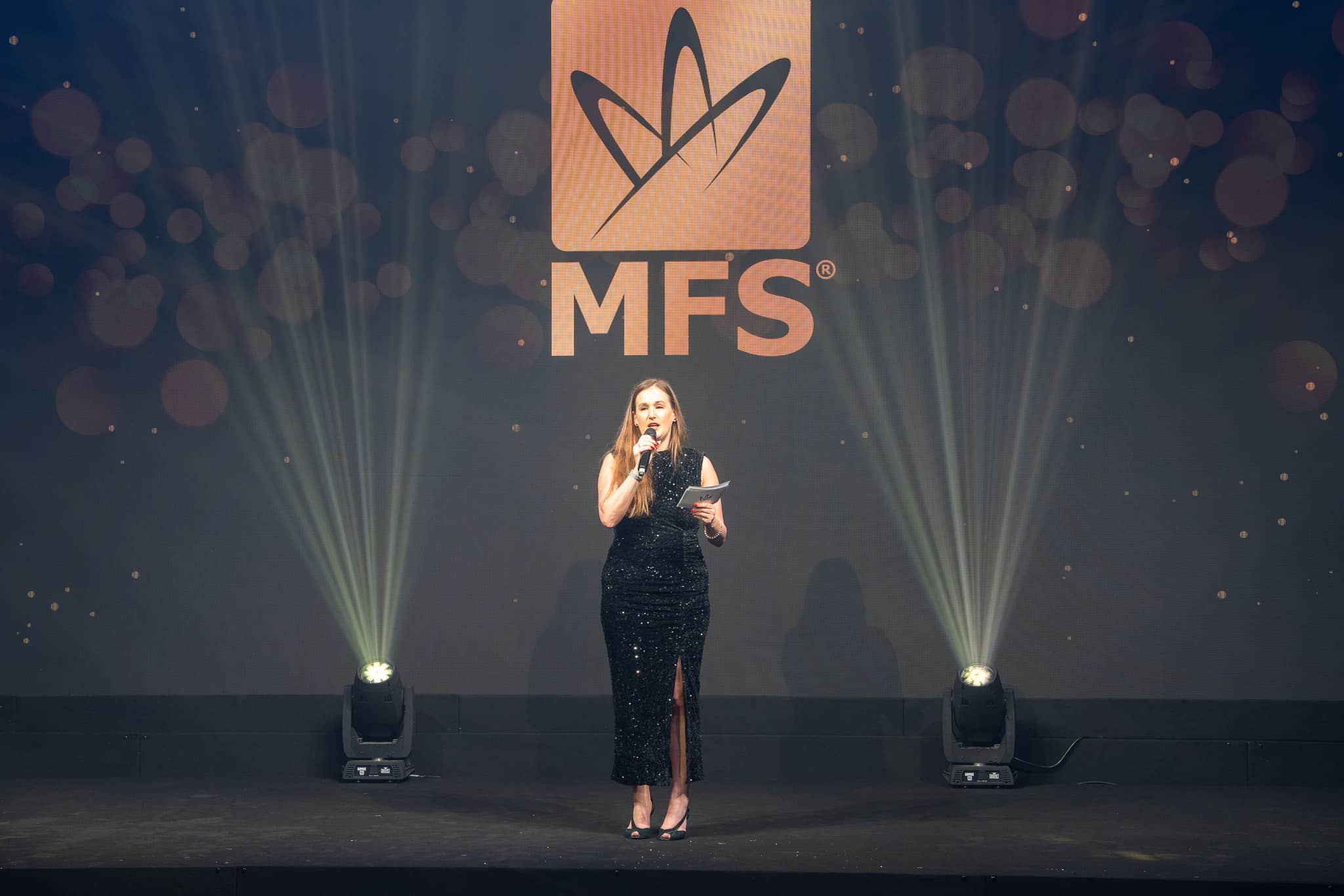 A presenter holding a microphone under the MFS screen logo with white stage beams