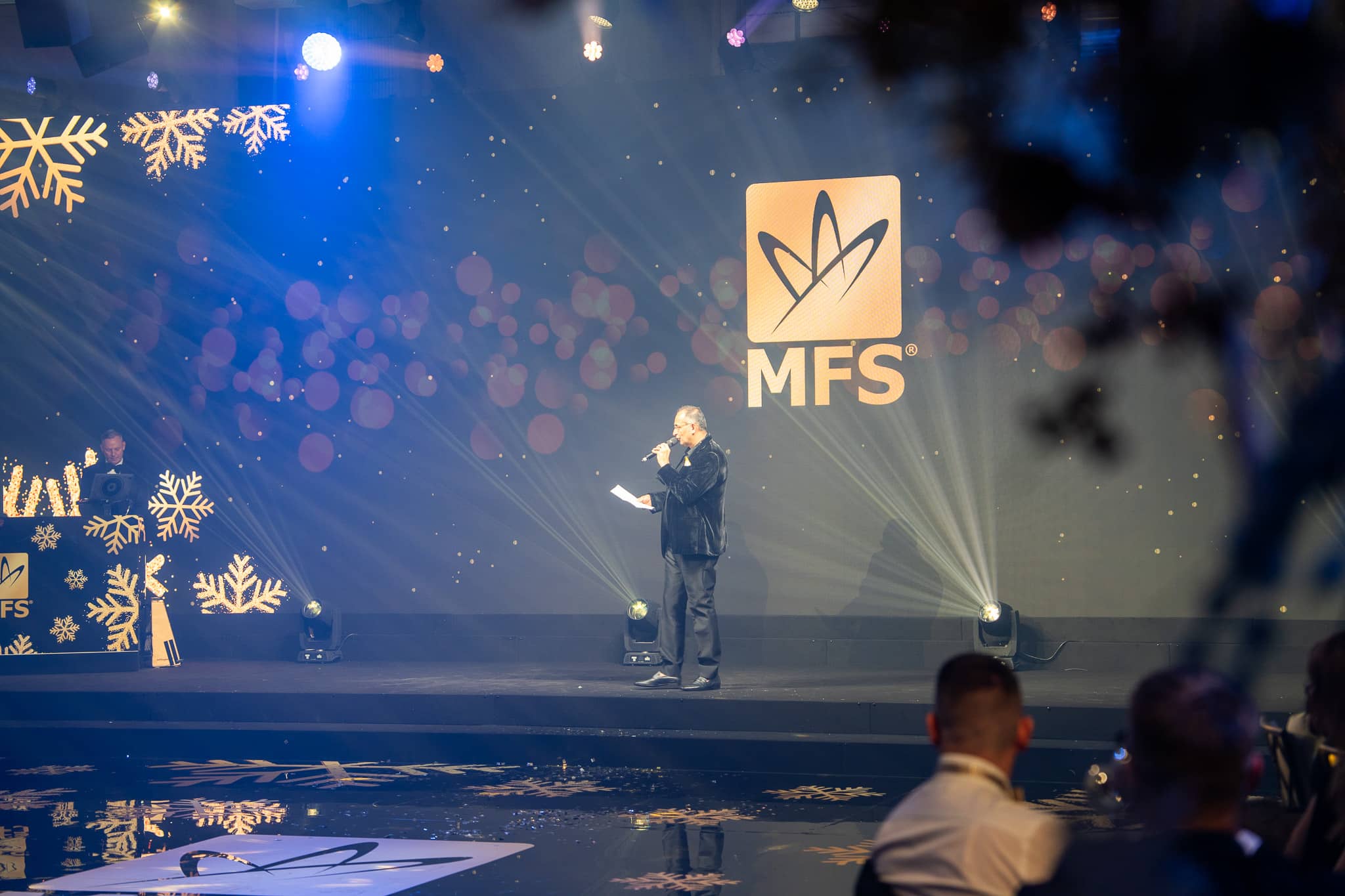 A presenter speaks beneath the gold MFS logo on the LED screen at The Peninsula London