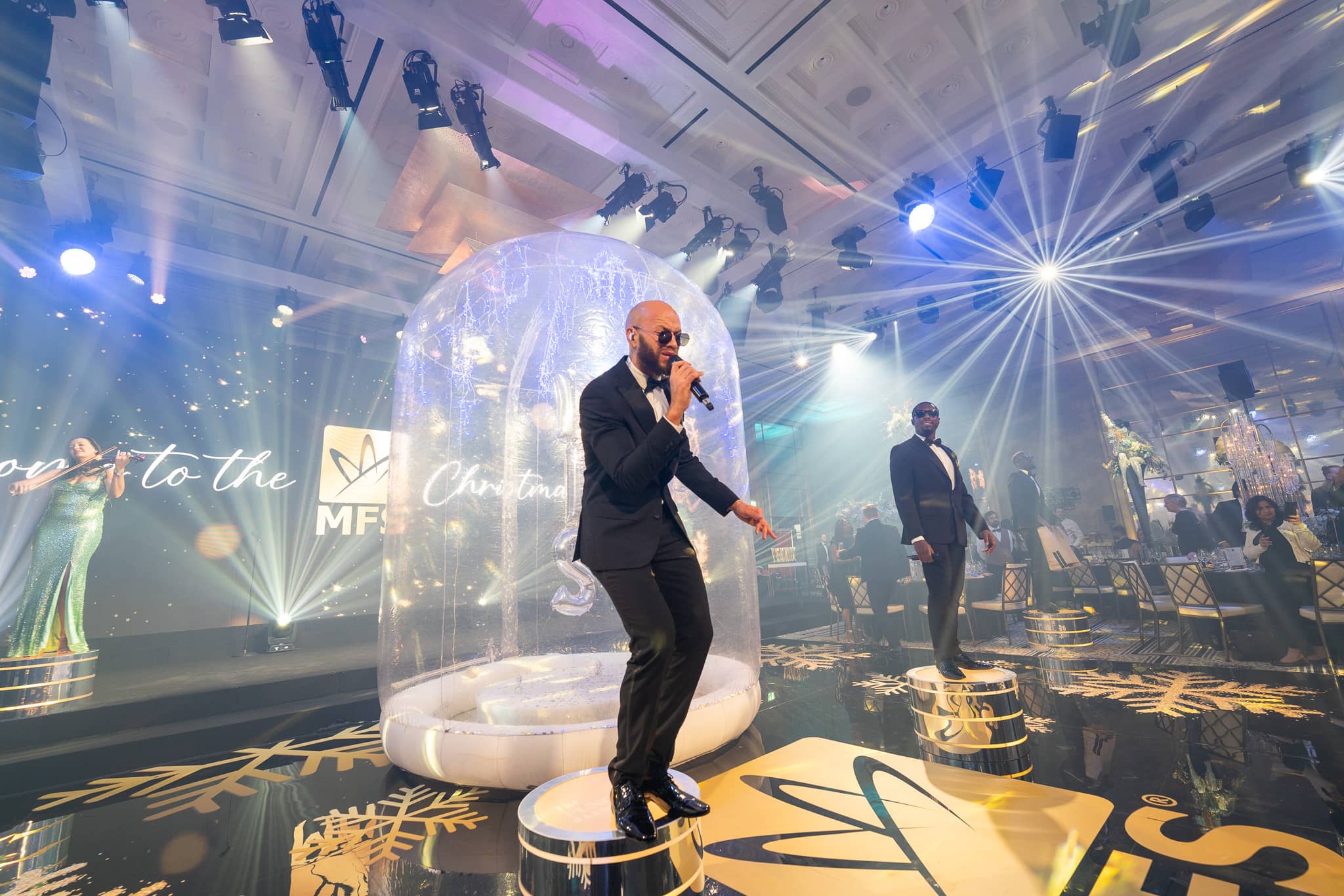 A vocalist with a handheld microphone stands on the gold MFS floor graphic beside the snow globe