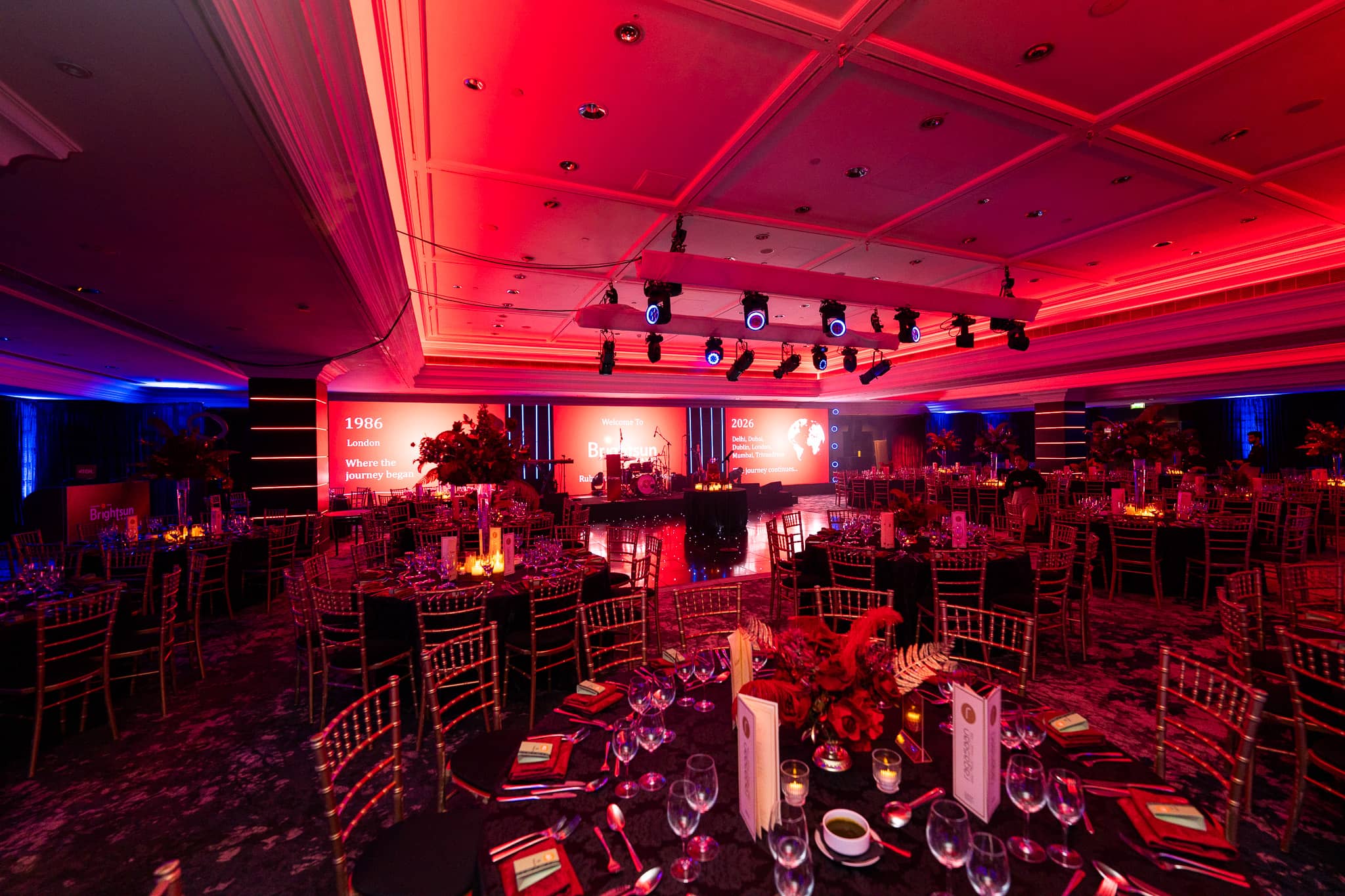 A long view through the ballroom towards the lit stage, with red ceiling uplighting, at Brightsun Travel’s 40th anniversary at InterContinental London Park Lane.