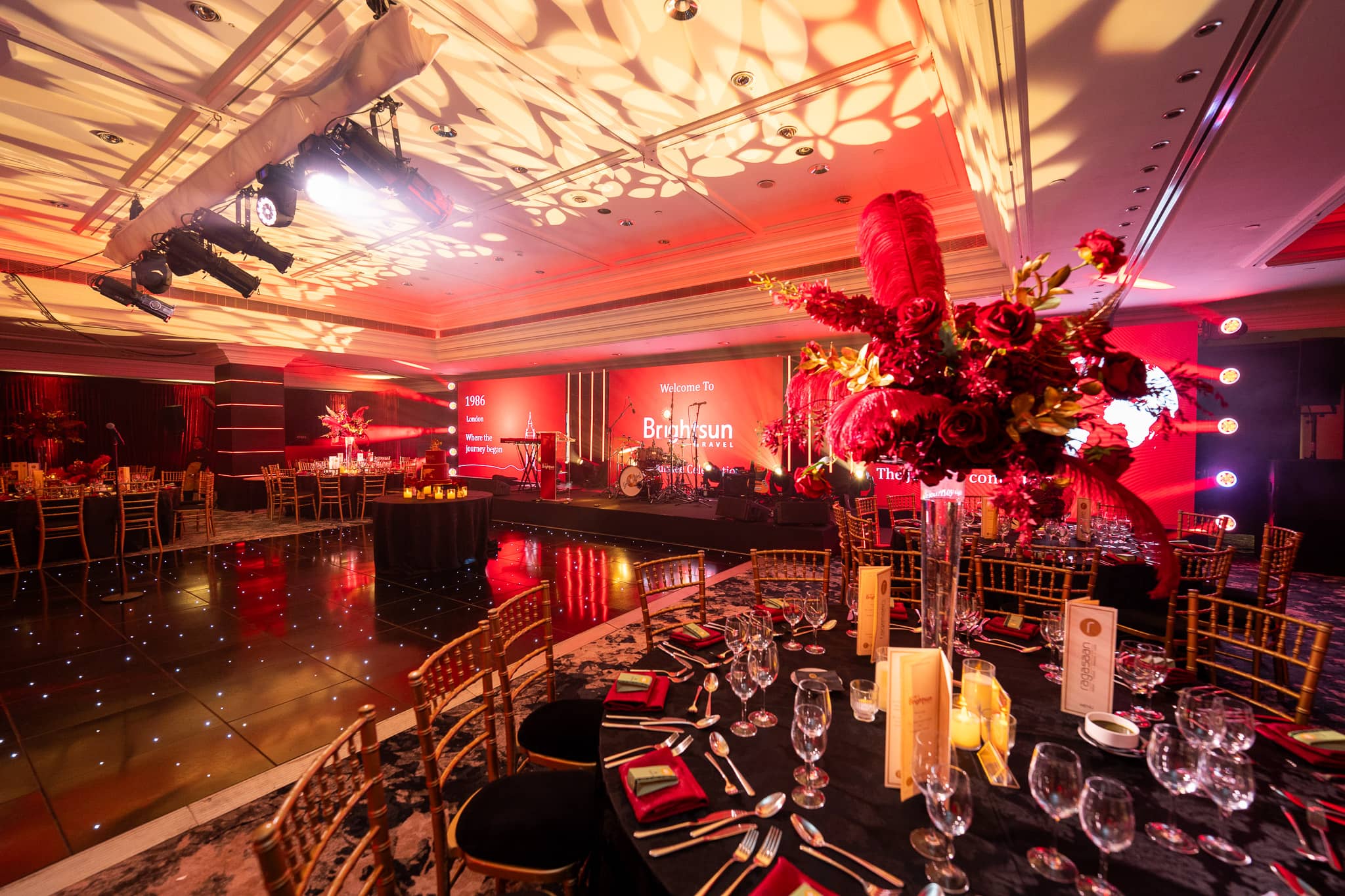 Dinner tables and red florals beneath patterned overhead lighting, at Brightsun Travel’s 40th anniversary at InterContinental London Park Lane.