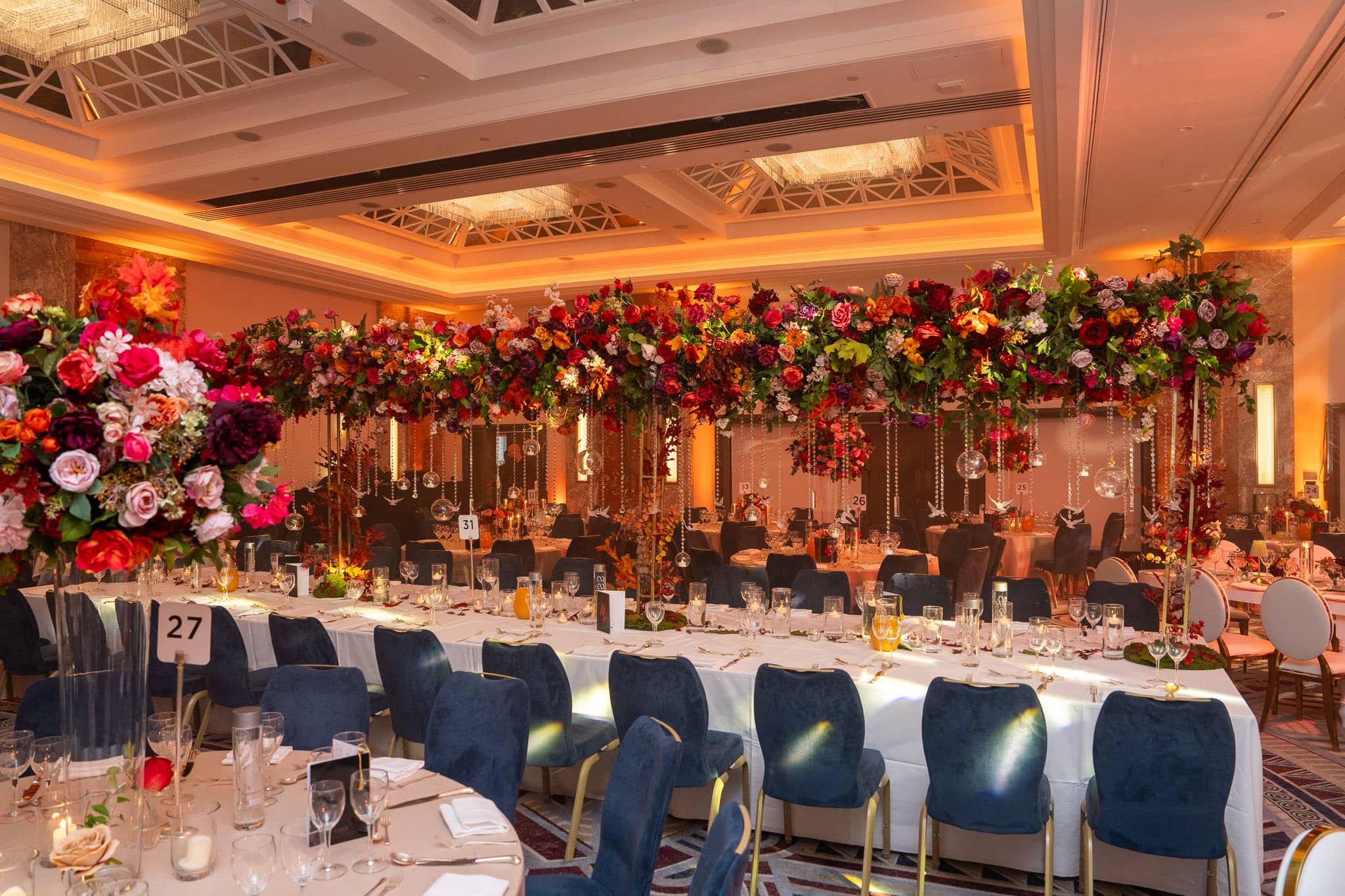 Long banquet tables and blue chairs beneath red floral canopies in the warmly lit ballroom.