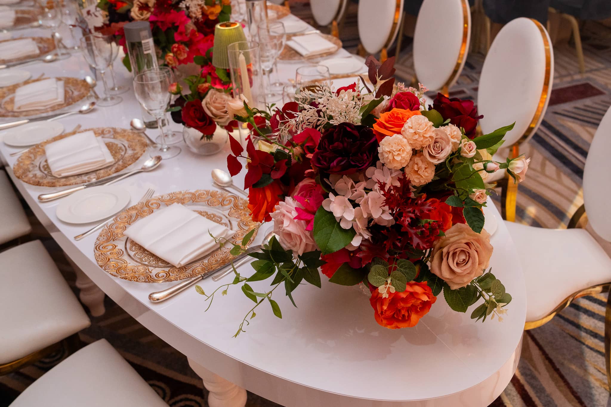 White curved table with gold-rimmed place settings and red and pink flowers at Pooja and Gary’s wedding.