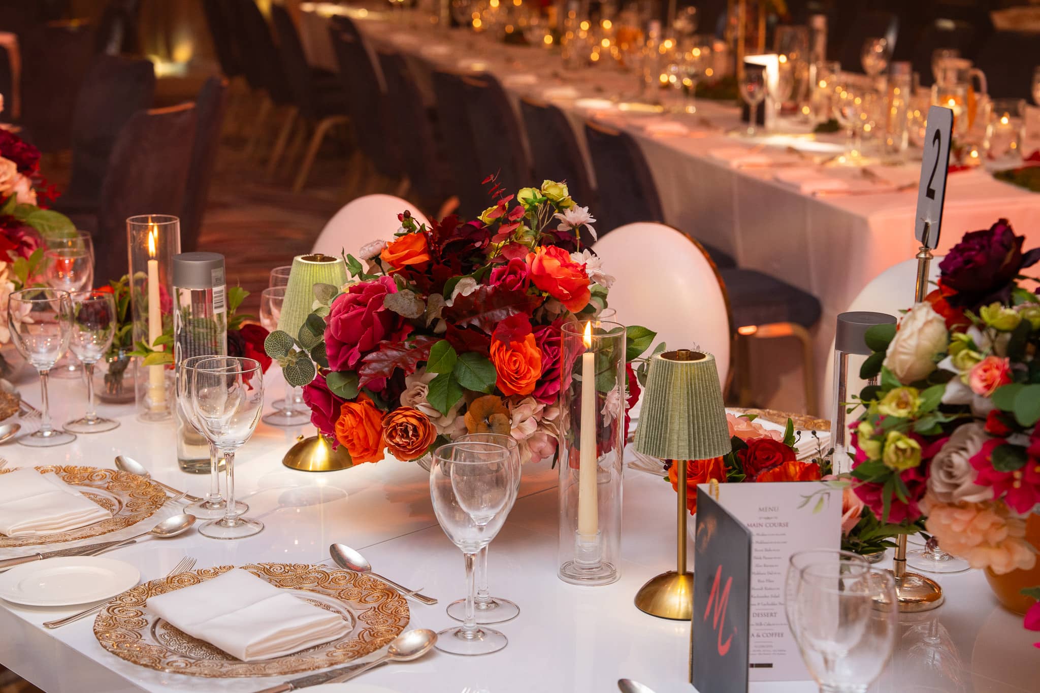 A place setting on a white bespoke table with a small green lamp and red floral arrangement.