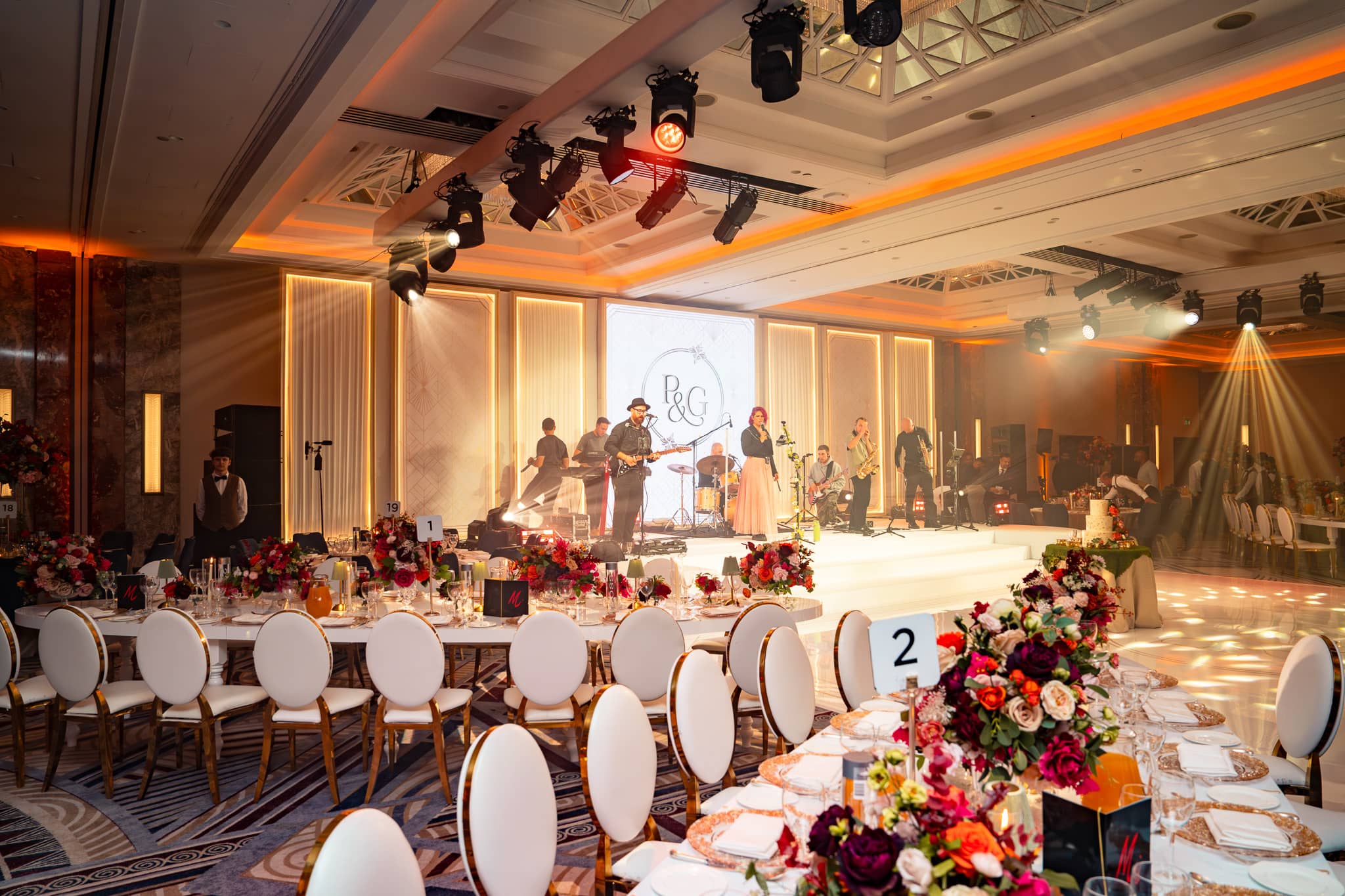 Performers on the white stage beneath overhead lights, with the Pooja and Gary monogram on the LED screen.