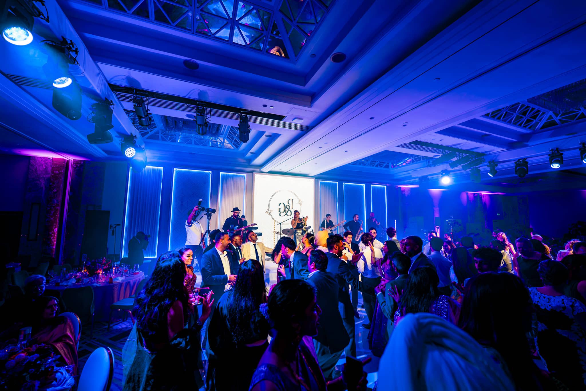A busy dancefloor under blue lighting with the illuminated stage and central LED screen in the background.