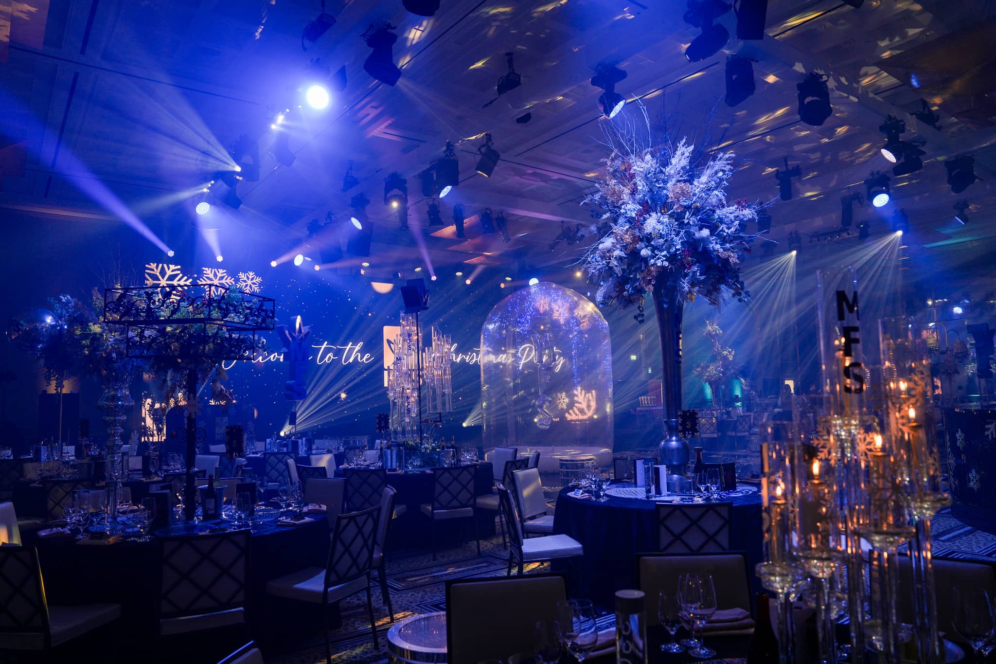 Blue-lit ballroom with tall floral centrepieces and the MFS Christmas party screen at The Peninsula London