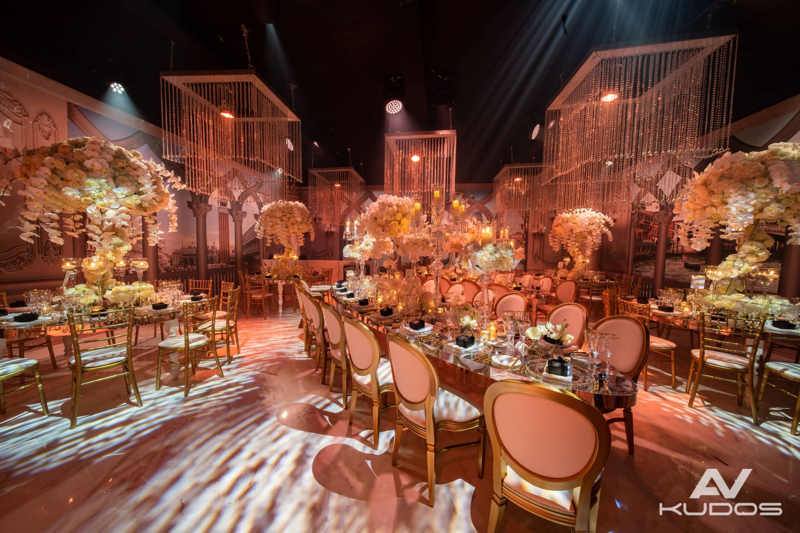 Wide view of the wedding ballroom at Claridge’s, with long dining tables, floral centrepieces, suspended chandeliers and patterned lighting across the floor