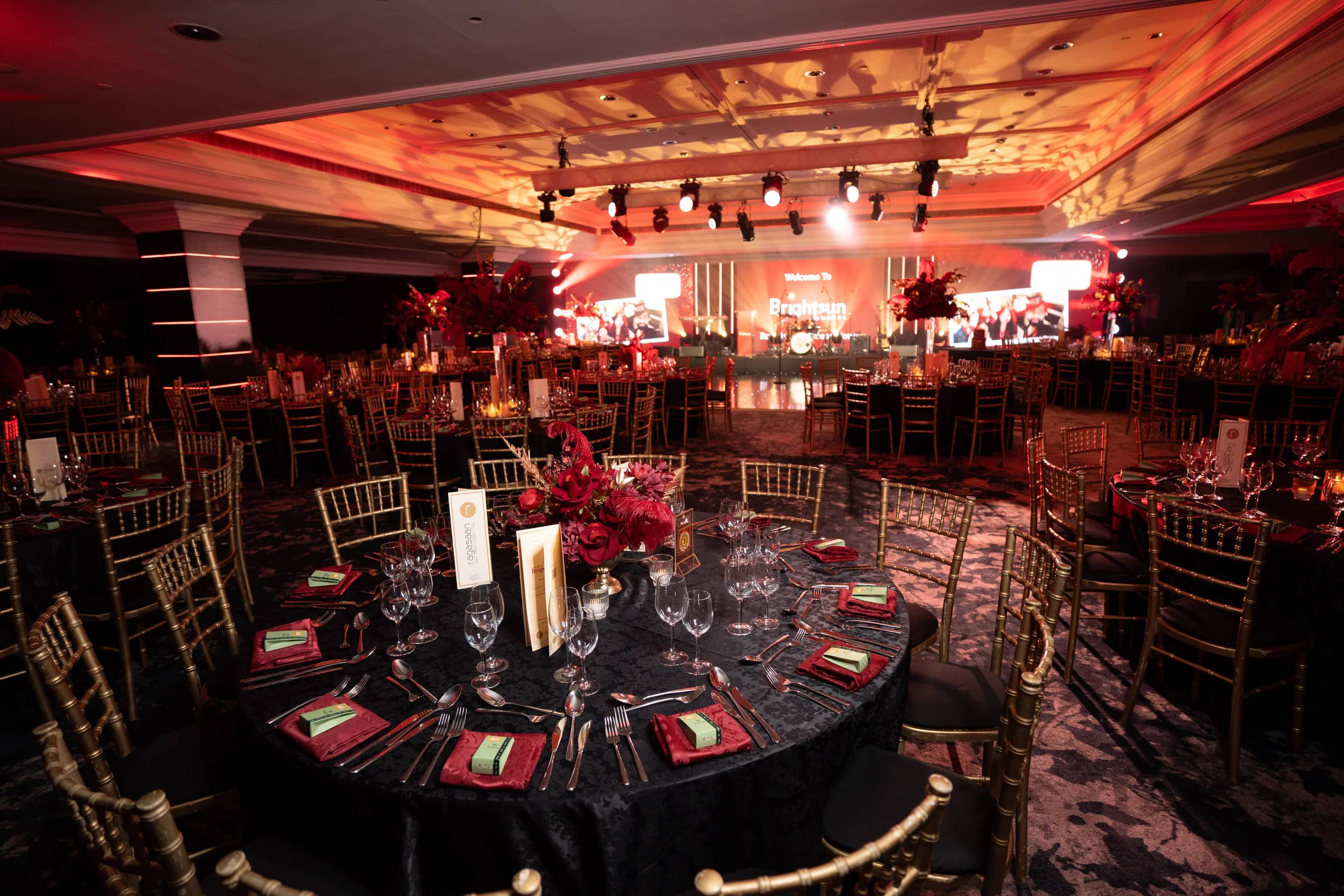 A dressed dinner table looking towards the anniversary stage beneath gold ceiling patterns and ruby lighting, at Brightsun Travel’s 40th anniversary at InterContinental London Park Lane.